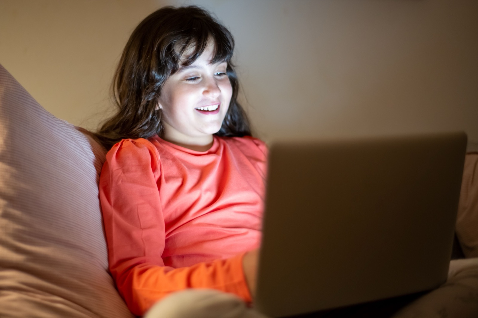 A girl smiling at a laptop screen, its glow lighting her face in a dim room