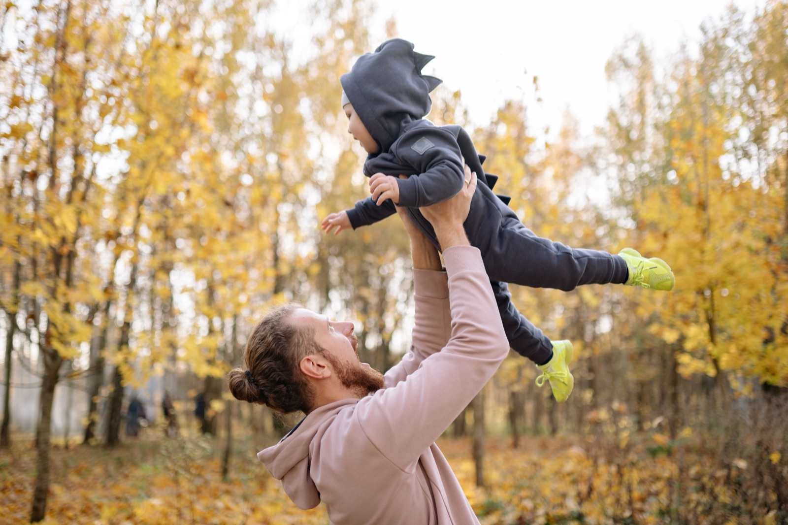 A father lifting his baby, dressed in a dinosaur onesie, into the air in an autumn park