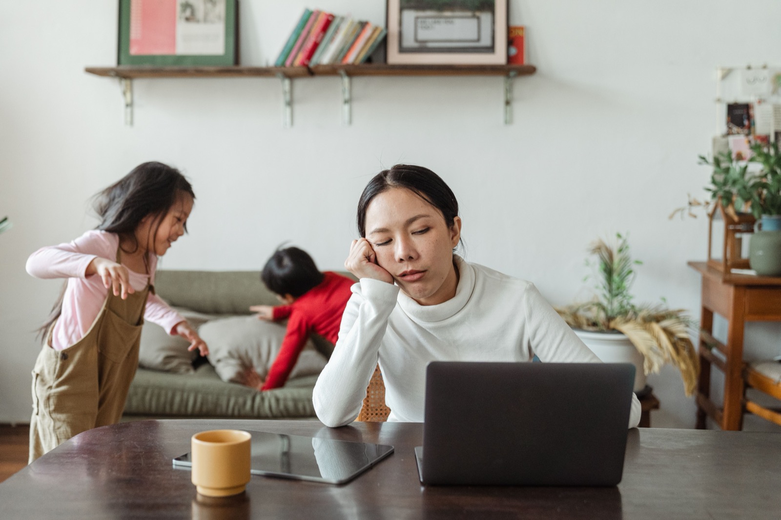 A tired parent rests her chin in her hand at the table in front of a laptop while two children clamber over the sofa behind her